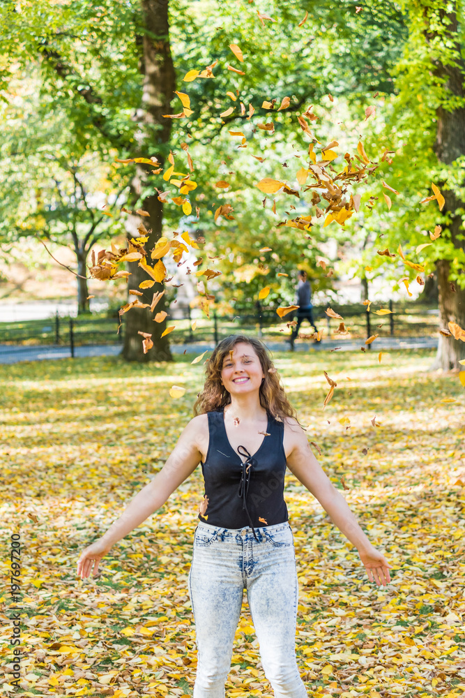 Manhattan New York City NYC Central park, young hipster millennial woman standing, throwing many fallen leaves up in air in autumn fall season with yellow foliage Manhattan New York City NYC Central park, young hipster millennial woman standing, throwing many fallen leaves up in air in autumn fall season with yellow foliage