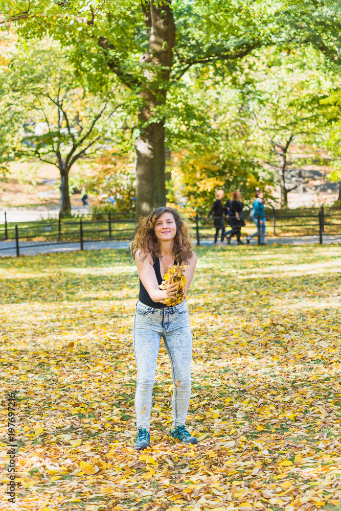 Manhattan New York City NYC Central park, young hipster millennial woman standing, throwing many fallen leaves up in air in autumn fall season with yellow foliage Manhattan New York City NYC Central park, young hipster millennial woman standing, throwing many fallen leaves up in air in autumn fall season with yellow foliage