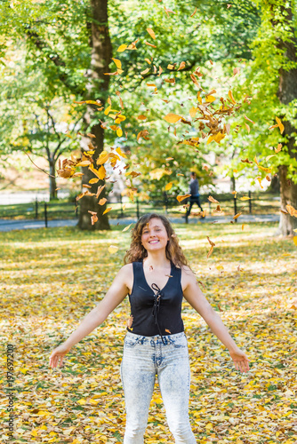 Manhattan New York City NYC Central park, young hipster millennial woman standing, throwing many fallen leaves up in air in autumn fall season with yellow foliage