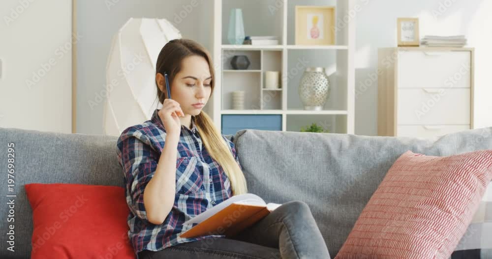 Caucasian young pretty woman sitting on the sofa with pillows and doing notes in the notebook or diary in the cozy living room. Indoors