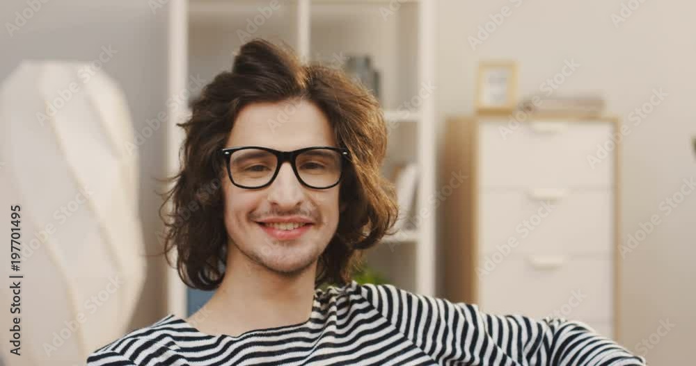 Close up of the young Caucasian man with a long hair and in glasses looking at the side then turning his head to the camera in the light living room. Portrait. Indoor Close up of the young Caucasian man with a long hair and in glasses looking at the side then turning his head to the camera in the light living room. Portrait. Indoor