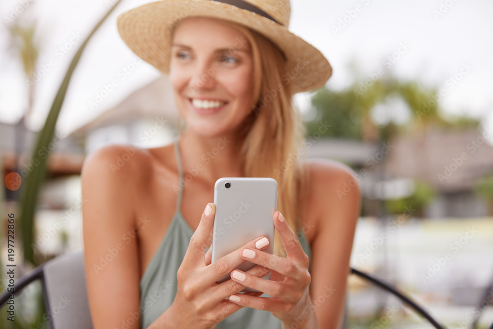 Fototapeta premium Cheerful relaxed woman uses smart phone for chat with friends, sits in modern coffee shop or terrace cafe. Pretty female reads good news on internet website, enjoys summer rest. Focus on cell phone