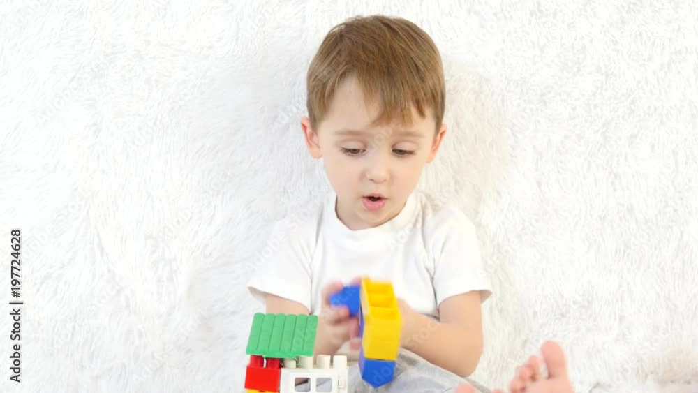 The little boy builds a toy house of colored blocks, sitting on a white background, close-up. Child and toys.