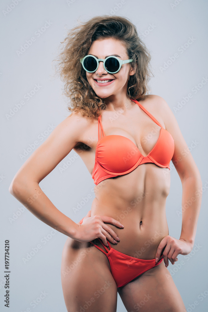 Fototapeta premium Young woman in sunglasses with beautiful slim perfect body in bikini isolated gray background