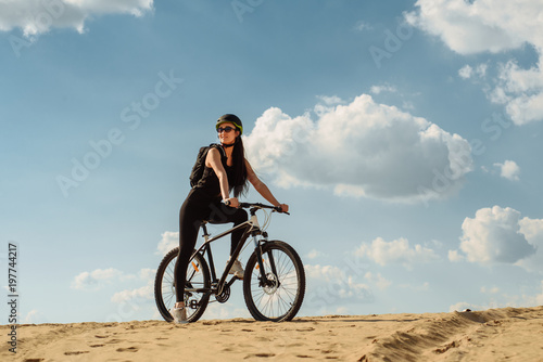Cyclist woman in helmet and sport wear with mountain bike on the slope under blue sky. Athlete girl is running with her extreme bike outdoors.