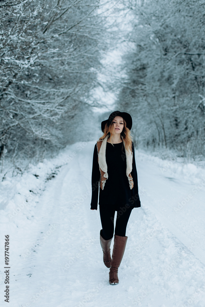a young stylish woman in a black hat is walking in a winter snowy forest