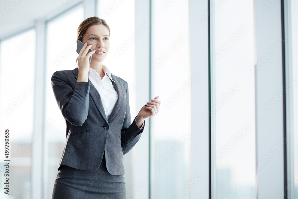 Young elegant businesswoman standing by office window and talking to one of clients by smartphone Young elegant businesswoman standing by office window and talking to one of clients by smartphone