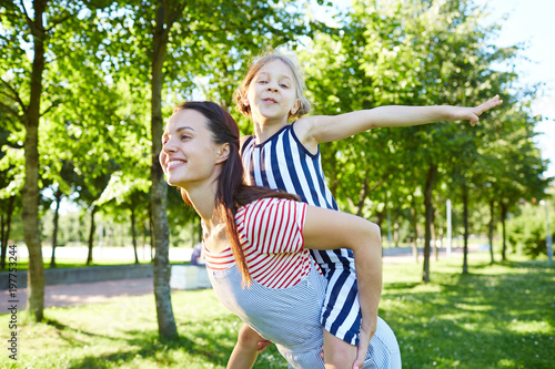 Happy female in casualwear holding little girl with outstreched arms on her back in park