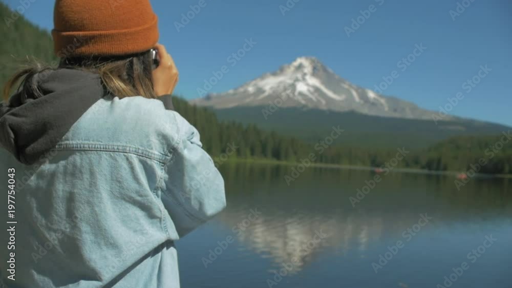 Panning over the shoulder shot of a young woman taking a picture of a mountain with her vintage camera by the lake shore on a sunny day Panning over the shoulder shot of a young woman taking a picture of a mountain with her vintage camera by the lake shore on a sunny day