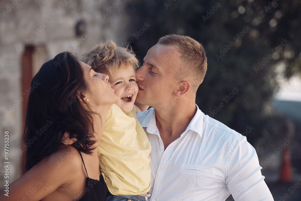 Fototapeta premium Happy family spend time together, urban background. Parents kissing son in cheeks, happy boy with smiling face. Mother and father love their child. Happy family concept. Parenthood, hugs, love, kiss.