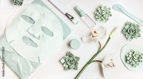 Skin care flat lay with facial sheet mask, mist spray bottle , succulents and orchid flowers on white desktop background, top view. Beauty spa and wellness concept photo
