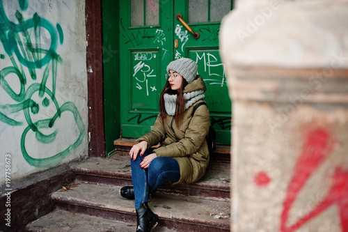 Portrait of brunette girl in gray scarf and hat, glasses sitting against urban entrance.