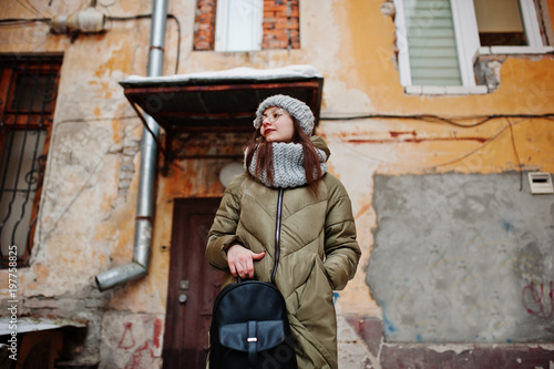 Portrait of brunette girl in gray scarf and hat, glasses at cold weather against orange wall of old house.