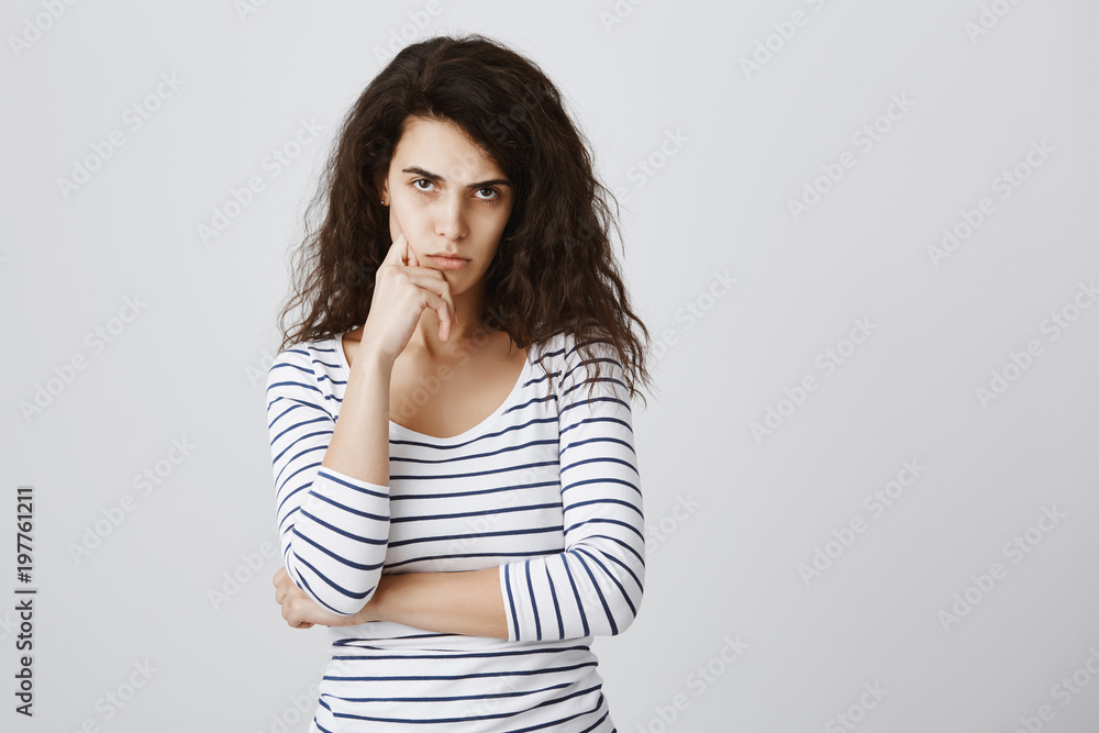 Studio portrait of offended cute curly-haired female looking from under forehead, holding hand on chin, being bothered and annoyed, thinking about revenge after insult, standing over gray wall Studio portrait of offended cute curly-haired female looking from under forehead, holding hand on chin, being bothered and annoyed, thinking about revenge after insult, standing over gray wall