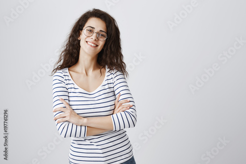 Active trendy young woman ready to take care of work. Beautiful smart curly-haired girl in glasses standing with raised chin and crossed hands, smiling broadly, being confident in her decisions