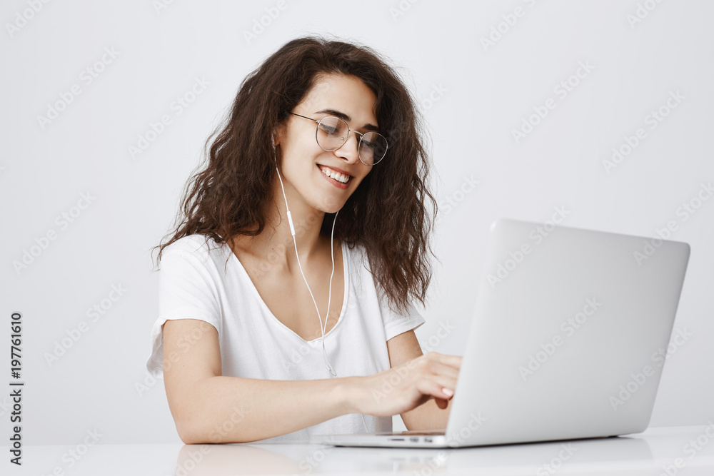 Typing message to answer on tricky question from friend. Pleased happy young woman working with laptop, smiling broadly, enjoying great sound while listening music in earphones, browsing in net Typing message to answer on tricky question from friend. Pleased happy young woman working with laptop, smiling broadly, enjoying great sound while listening music in earphones, browsing in net
