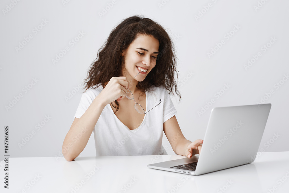 Having great day, enjoying using new laptop. Indoor shot of pleased carefree european woman biting rim of glasses and smiling broadly while looking at screen and typing message in notebook Having great day, enjoying using new laptop. Indoor shot of pleased carefree european woman biting rim of glasses and smiling broadly while looking at screen and typing message in notebook