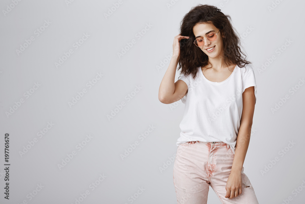 Living fashionable and modern life. Portrait of stylish urban curly-haired woman in trendy sunglasses and pink trousers looking down and smiling broadly while posing during advertising campaign Living fashionable and modern life. Portrait of stylish urban curly-haired woman in trendy sunglasses and pink trousers looking down and smiling broadly while posing during advertising campaign