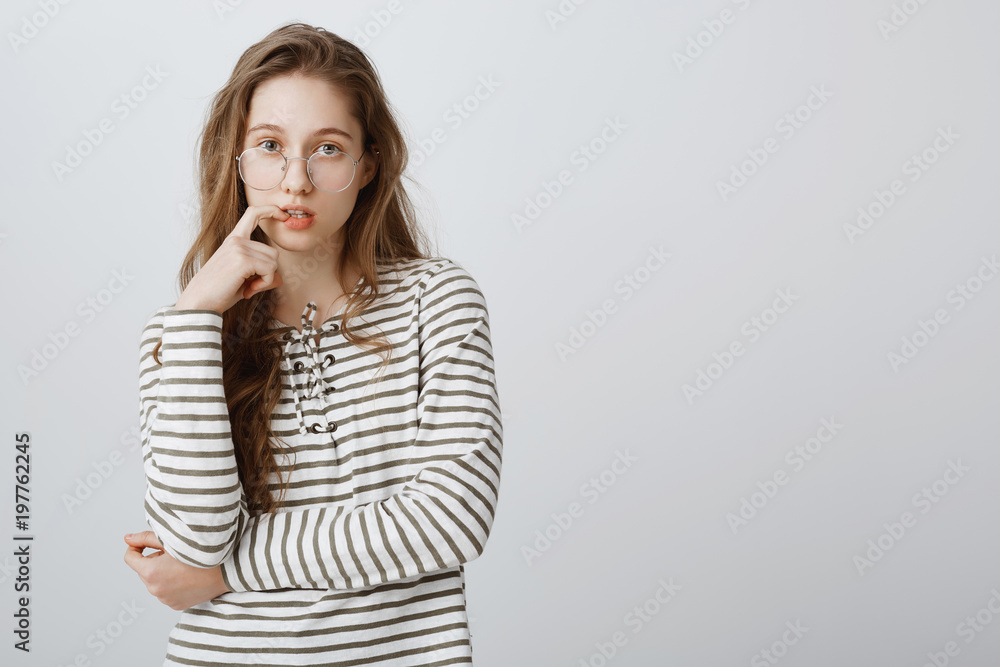 Good-looking young caucasian female in trendy glasses gazing at camera, biting finger while thinking or being interested in lection she attending, standing over gray background, smart and cute Good-looking young caucasian female in trendy glasses gazing at camera, biting finger while thinking or being interested in lection she attending, standing over gray background, smart and cute