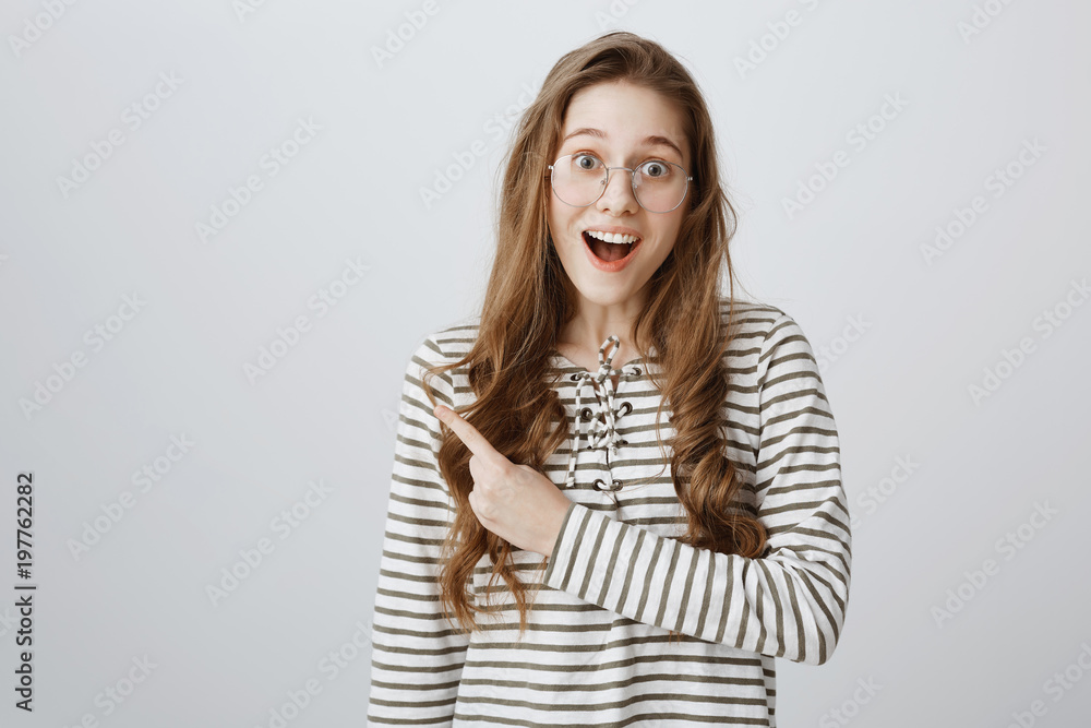 Studio portrait of attractive happy young woman in stylish glasses smiling with teeth from excitement and pointing left with forefinger, being thrilled of hearing positive news over gray background Studio portrait of attractive happy young woman in stylish glasses smiling with teeth from excitement and pointing left with forefinger, being thrilled of hearing positive news over gray background