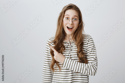 Studio portrait of attractive happy young woman in stylish glasses smiling with teeth from excitement and pointing left with forefinger, being thrilled of hearing positive news over gray background © Cookie Studio