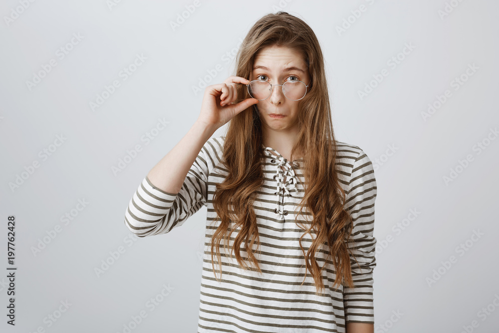 Studio shot of impressed young girl with blond hair, taking off glasses and looking from under forehead, pouting, being amazed and interested with great suggestion, curious over gray background Studio shot of impressed young girl with blond hair, taking off glasses and looking from under forehead, pouting, being amazed and interested with great suggestion, curious over gray background