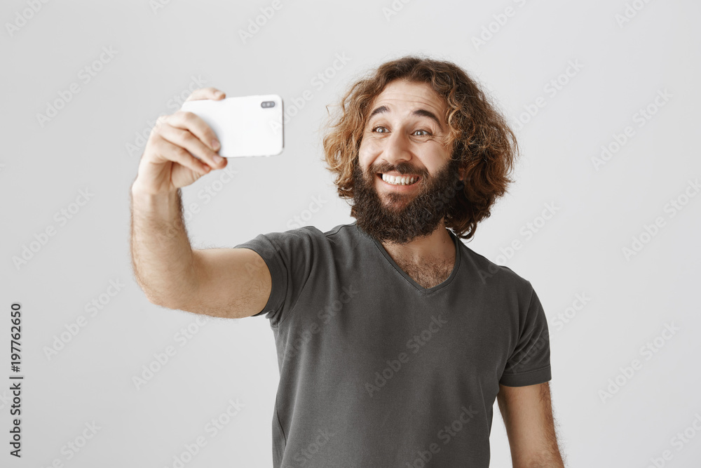 Guy takes selfie near new sportcar. Studio shot of wealthy good-looking eastern man with beard and curly hair making photo of himself with smartphone, smiling happily while standing over gray wall Guy takes selfie near new sportcar. Studio shot of wealthy good-looking eastern man with beard and curly hair making photo of himself with smartphone, smiling happily while standing over gray wall
