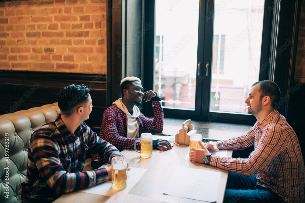 Multiracial friends talking and drinking beer and clinking glasses at pub