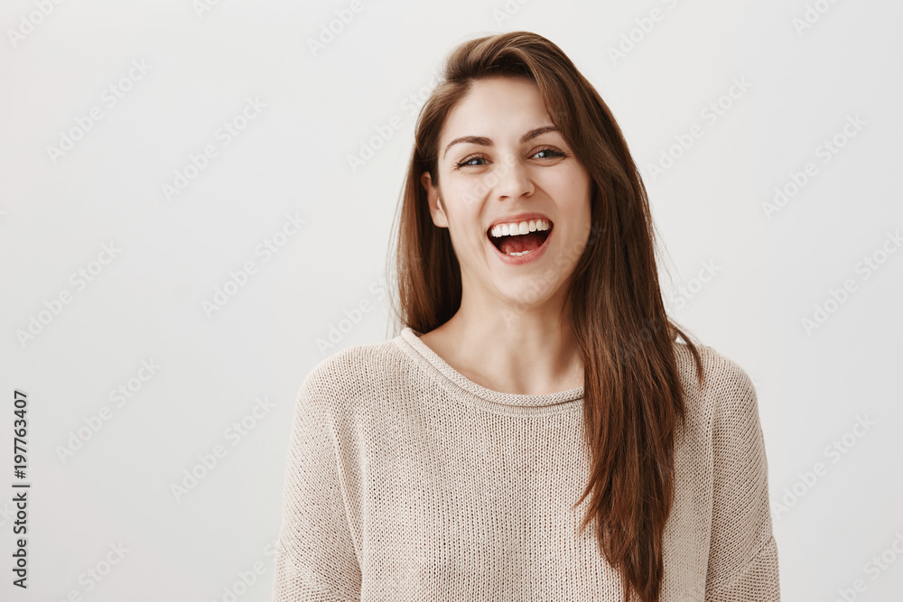 Indoor portrait of attractive ordinary european woman with long brown hair laughing out loud while gazing at camera, being in great mood, chuckling over video in network, standing against gray wall Indoor portrait of attractive ordinary european woman with long brown hair laughing out loud while gazing at camera, being in great mood, chuckling over video in network, standing against gray wall