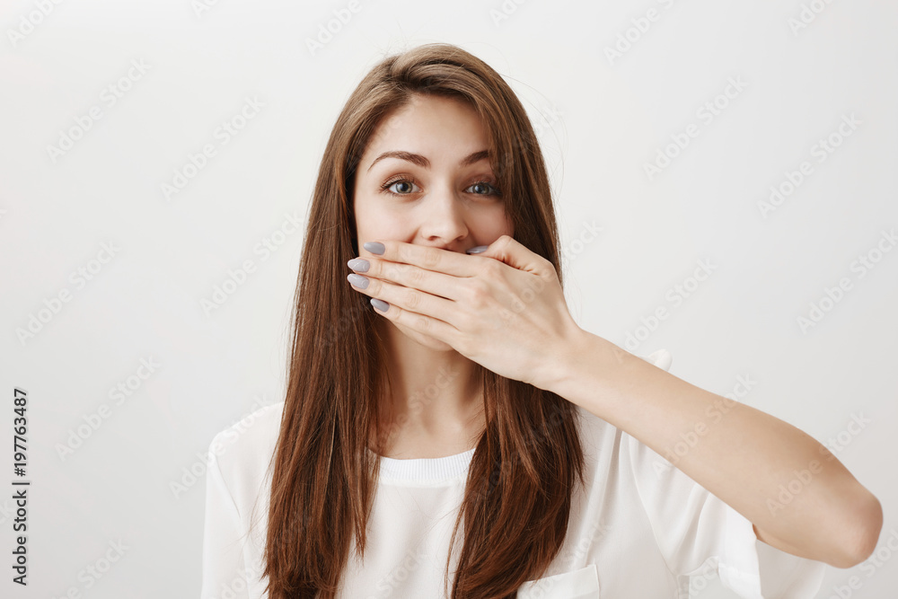 My mouth is shut of key. Portrait of gorgeous young european female covering mouth with palm and peeking at camera with natural expression, standing against gray background keeping secrets