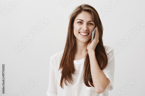 Let me finish this call. Studio shot of charming friendly girl with brown hair talking on smartphone and smiling broadly, gazing at camera, having conversation with best friend over gray background