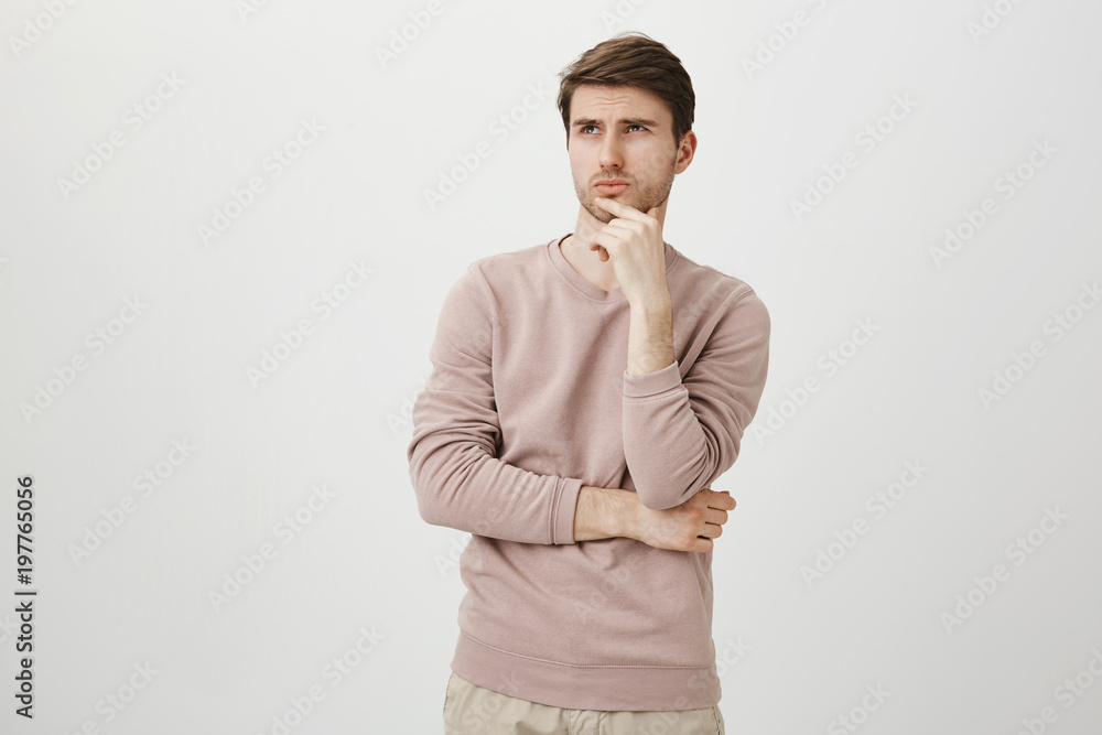 Portrait of attractive young man with bristle looking up while touching chin and thinking about something, standing over gray background in trendy pullover. Maybe I should skip practice today Portrait of attractive young man with bristle looking up while touching chin and thinking about something, standing over gray background in trendy pullover. Maybe I should skip practice today