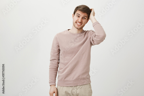 Indoor shot of puzzled and confused man with bristle scratching head and showing teeth while looking at camera and standing over gray background. Husband forgot to throw garbage and now says sorry