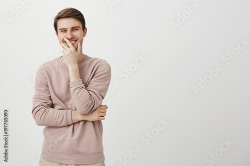 Portrait of positive charming european male covering mouth with hand while laughing and looking aside, standing over gray background. Guy is touched with sense of humour of girl he likes