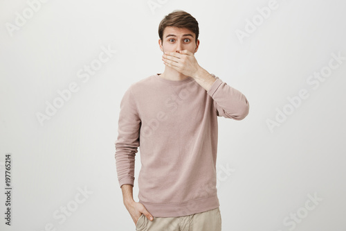 Portrait of surprised caucasian male lifting eyebrows and looking with widened eyes at camera while covering mouth with hand, standing against gray background. Secret and emotions concept.