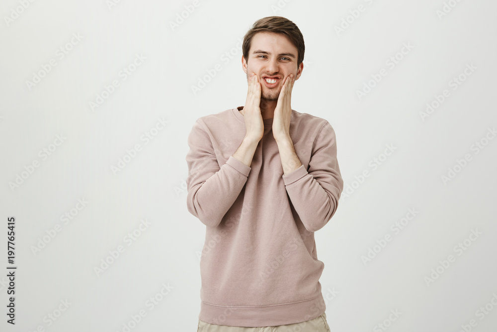 Portrait of funny handsome caucasian male squeezing cheeks with both hands and squinting at camera, standing against gray background. Guy looks in mirror and thinking that it is time to shave Portrait of funny handsome caucasian male squeezing cheeks with both hands and squinting at camera, standing against gray background. Guy looks in mirror and thinking that it is time to shave