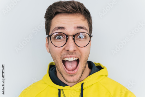 Closeup portrait of amazed young Caucasian man wearing trendy glasses and hoodie, keeping mouth wide open, looking shocked isolated over white studio background. People, lifestyle and emotion concept.