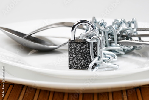 cutlery - spoon, fork and knife wrapped with chain and locked on padlock on white plate, diet concept