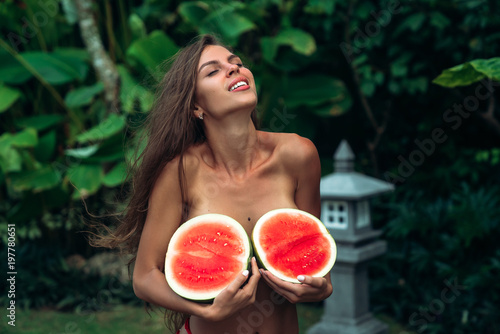 close up portrait of cute smiling brunette girl with a sporty body holds watermelons in her hands and covers her breasts. Photoshoot of a beautiful model with fruits near green palms on background in