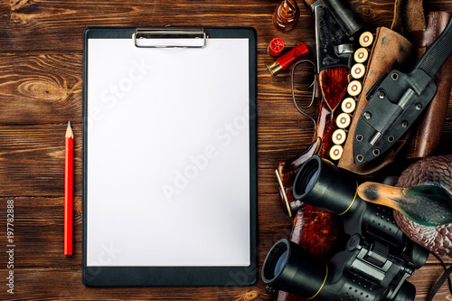 Blank clipboard and pen with hunting equipment on the wooden background.