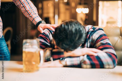 Young man in casual clothes is sleeping near the mug of beer on a table in pub, another man is waking him up. Get drunk man.