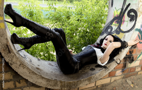 Gothic girl with dark hair and tight boots is lying down in the round window of the old building