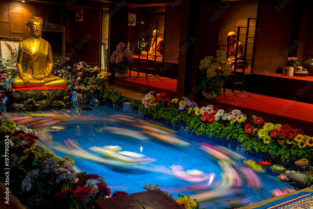 Fototapeta Colorful flowers floating under the golden statue of Buddha in Buddhist monastery. Buddha statue above the pool with moving water lilies.