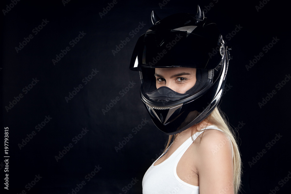 Beautiful self determined young European woman motorcyclist wearing white tank top and protective black helmet posing at blank studio wall with copy space for your text or promotional information