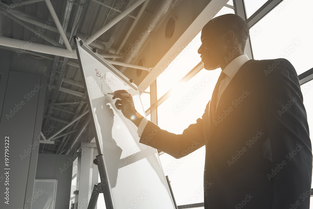 Low angle of african man in suit writing with marker on board. Sun is shining into large window