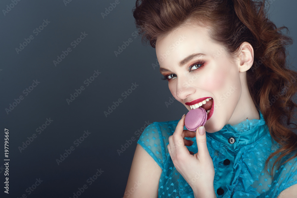 Close up portrait of a beautiful model with creative hairstyle and colourful make up eating violet macaroon and smiling. Isolated on blue background. Sweet tooth concept. Text space Close up portrait of a beautiful model with creative hairstyle and colourful make up eating violet macaroon and smiling. Isolated on blue background. Sweet tooth concept. Text space