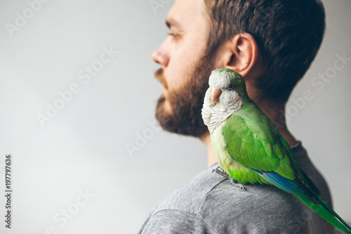 Young beard hipster man at home with his favorite pet on shoulder - green monk parrot. Monk parakeet is looking at camera with curiosity. Copy- space area for your advertisement text