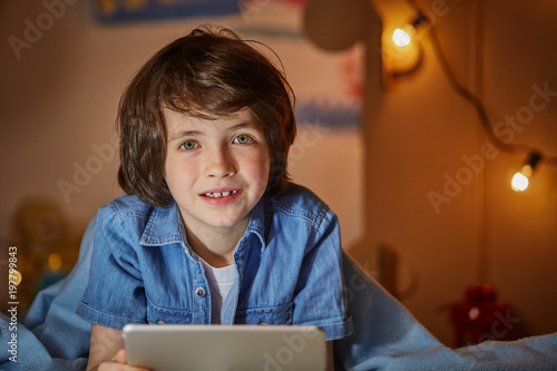 Waist up portrait of pleased kid lying under plaid and using clipboard. He is looking at camera with happiness and joyfulness