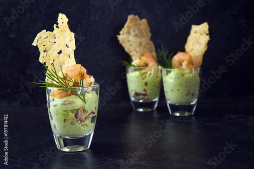 avocado cream or guacamole with shrimps and cheese crisp in a glass, appetizer or party snack on a dark slate background with copy space