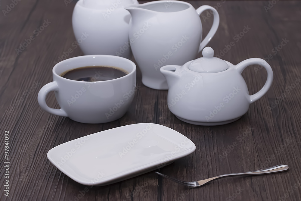 A white cup of coffee and other white utensils on brown wooden table. Breakfast. A white cup of coffee and other white utensils on brown wooden table. Breakfast.